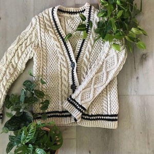 May include: Cream-coloured cable knit cardigan with black trim and buttons. The sweater features a V-neck, long sleeves, and a textured knit pattern. The cuffs and hem have black stripes. The cardigan is displayed on a light wooden surface.