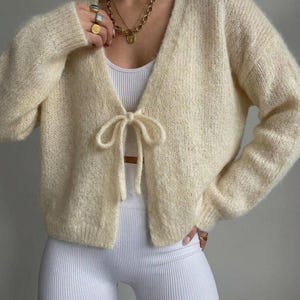 Op de afbeelding: Crèmekleurig vest met open voorkant en striksluiting. De zachte gebreide trui heeft lange mouwen en wordt gecombineerd met een witte tanktop en geribbelde legging. Een gouden ketting en ringen maken de look compleet. Mode kleding.