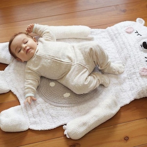 May include: A baby in a light-coloured onesie lies on a white, bear-shaped knitted rug. The rug has a grey circular centre and details like pink cheeks and closed eyes. The rug is on a wooden floor.