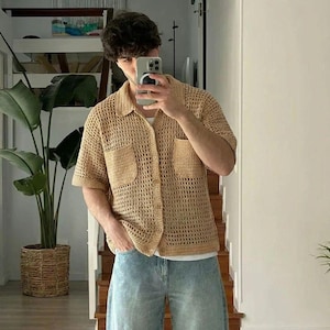 May include: A tan crochet shirt with short sleeves and two front pockets. The shirt is buttoned up and worn over a white undershirt. The person is wearing light blue denim jeans. The shirt has a collared neckline.