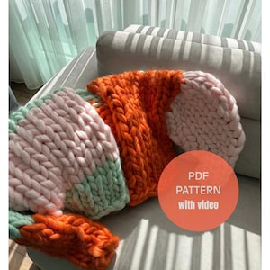 Puede incluir: Una manta de punto grueso en tonos naranja, rosa y verde menta descansa sobre un sofá gris. La manta está hecha de hilo grueso, con un patrón de puntada visible. Un gráfico redondo naranja con el texto "PDF PATTERN with video" es visible.