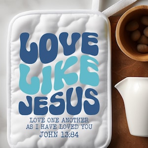 May include: White quilted pot holder with the text "LOVE LIKE JESUS" in blue and teal. The pot holder also has the text "LOVE ONE ANOTHER AS I HAVE LOVED YOU JOHN 13:34". A white loop is attached for hanging.