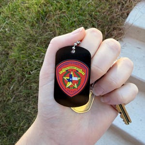 May include: Black dog tag with a silver ball chain, featuring the Texas Highway Patrol emblem. The emblem is a shield shape with red, gold, and blue accents, and the Texas star in the center. A gold key and tag are also visible.