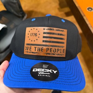 Peut inclure: Casquette de baseball noire et bleue avec un patch en cuir marron représentant le drapeau américain, l'année 1776 et le texte "WE THE PEOPLE". La casquette a une visière incurvée et la marque "DECKY Classic Original".