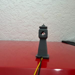 May include: A gray, 3D-printed miniature lighthouse with a diamond-shaped opening. The lighthouse sits on a red surface, with yellow and black wires extending from its base. The background is a textured white wall.