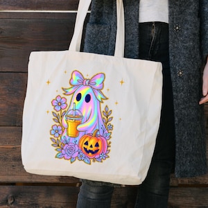 May include: A cream-colored tote bag featuring a colorful ghost design holding a drink, surrounded by flowers and a jack-o'-lantern. The ghost has a rainbow sheen, and the design includes a bow, stars, and floral elements, perfect for Halloween.