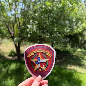 Op de afbeelding: Een schildvormige geborduurde patch met een rode achtergrond en een centrale gouden ster met een blauwe en witte vlag van Texas. De tekst luidt Department of Public Safety en Texas Highway Patrol met het woord Texas onderaan.