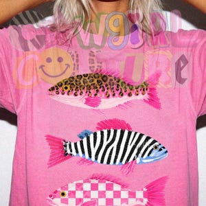 May include: Pink t-shirt featuring three stylized fish designs. One fish has a leopard print pattern, another has black and white stripes, and the third has a pink and white checkerboard pattern. The shirt also has the text "W91RL COUTURE".