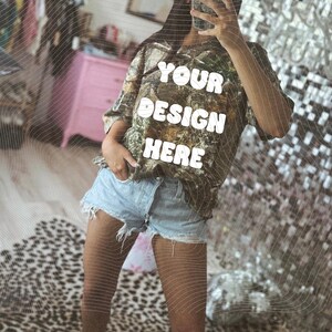 May include: A camouflage-patterned t-shirt with the text "YOUR DESIGN HERE" in white block letters. The person is wearing light blue denim shorts and white sneakers. A pink baseball cap is worn.