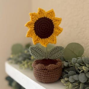 May include: A handcrafted crochet sunflower in a brown pot. The sunflower has yellow petals and a dark brown center. Two green leaves are below the flower. The pot is brown with a scalloped edge. The item is a decorative home accent.
