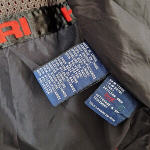 May include: Close-up of a black and red jacket interior with multiple fabric content and care instruction tags. The tags are blue with white text, detailing the materials and dry-clean-only instructions. The jacket has a mesh lining.
