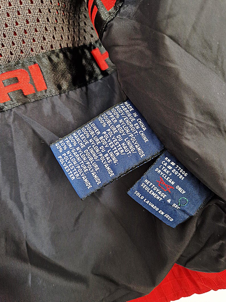 May include: Close-up of a black and red jacket interior with multiple fabric content and care instruction tags. The tags are blue with white text, detailing the materials and dry-clean-only instructions. The jacket has a mesh lining.