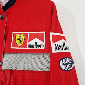 May include: Red Ferrari racing jacket with multiple sponsor patches, including Marlboro and Magneti Marelli. The jacket features a black and white checkered stripe across the chest and a Ferrari logo patch.