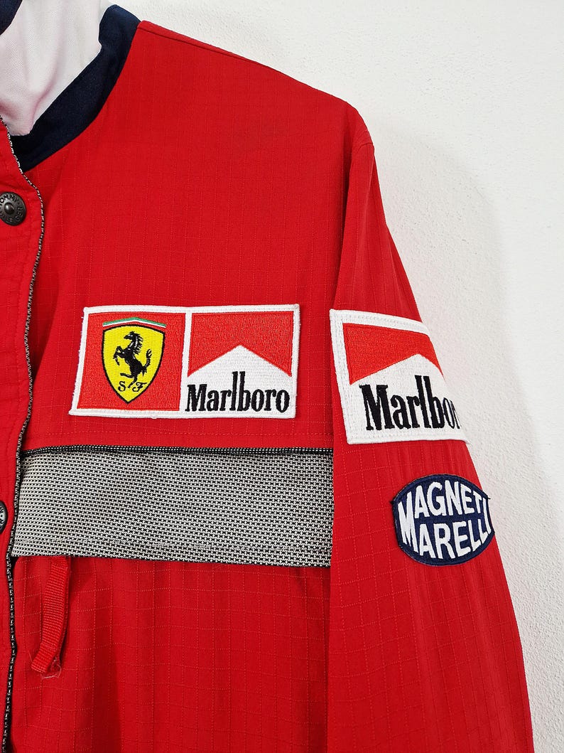 May include: Red Ferrari racing jacket with multiple sponsor patches, including Marlboro and Magneti Marelli. The jacket features a black and white checkered stripe across the chest and a Ferrari logo patch.