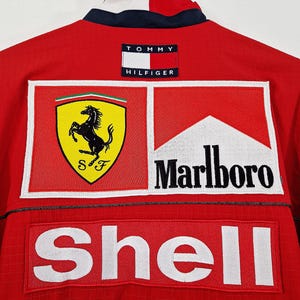 May include: Red Tommy Hilfiger jacket with Ferrari, Marlboro, and Shell logos. The jacket has a white and navy blue collar and a Tommy Hilfiger patch on the back. The jacket is made of a textured material.