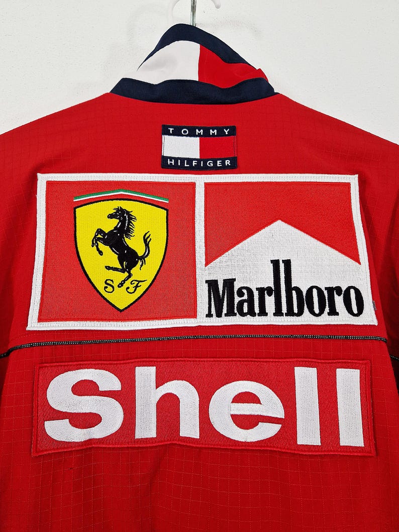 May include: Red Tommy Hilfiger jacket with Ferrari, Marlboro, and Shell logos. The jacket has a white and navy blue collar and a Tommy Hilfiger patch on the back. The jacket is made of a textured material.