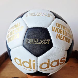 May include: A vintage Adidas Telstar soccer ball featuring a classic black and white pentagonal panel design. The ball is printed with gold text including "TELSTAR," "OFFICIAL WORLD CUP MEXICO," "DURLAST," and "adidas MADE IN FRANCE."
