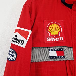 May include: Red racing jacket with multiple sponsor patches. Patches include "Marlboro", "Shell", and "Tommy Hilfiger". The jacket has a navy blue collar and a checkered pattern detail.