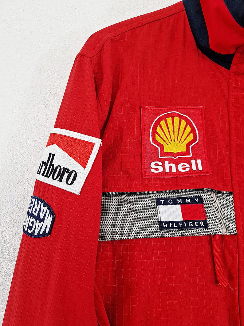 May include: Red racing jacket with multiple sponsor patches. Patches include "Marlboro", "Shell", and "Tommy Hilfiger". The jacket has a navy blue collar and a checkered pattern detail.