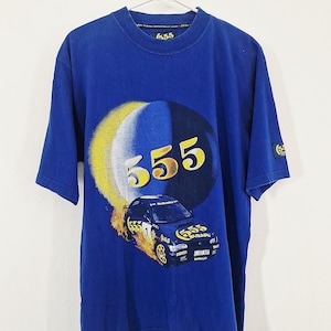May include: A royal blue short-sleeve t-shirt featuring a large graphic of a rally car with the number 555 in yellow and white. The design includes a circular yellow and blue background element, the text 555, and a small patch on the left sleeve.