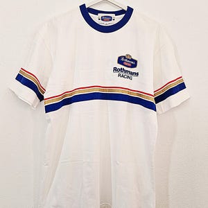 May include: A white short-sleeve t-shirt featuring a blue crew neckline and a horizontal stripe pattern in blue, gold, and red across the chest and sleeves. The chest displays a Rothmans Racing logo with the text Rothmans RACING below it.