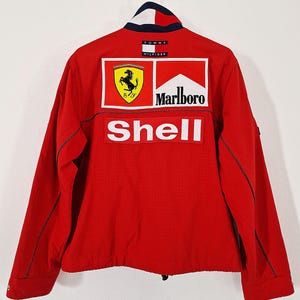 May include: Red jacket with a white, blue, and red collar. The back of the jacket features the Ferrari logo, the Marlboro logo, and the Shell logo. The jacket is a Tommy Hilfiger design.