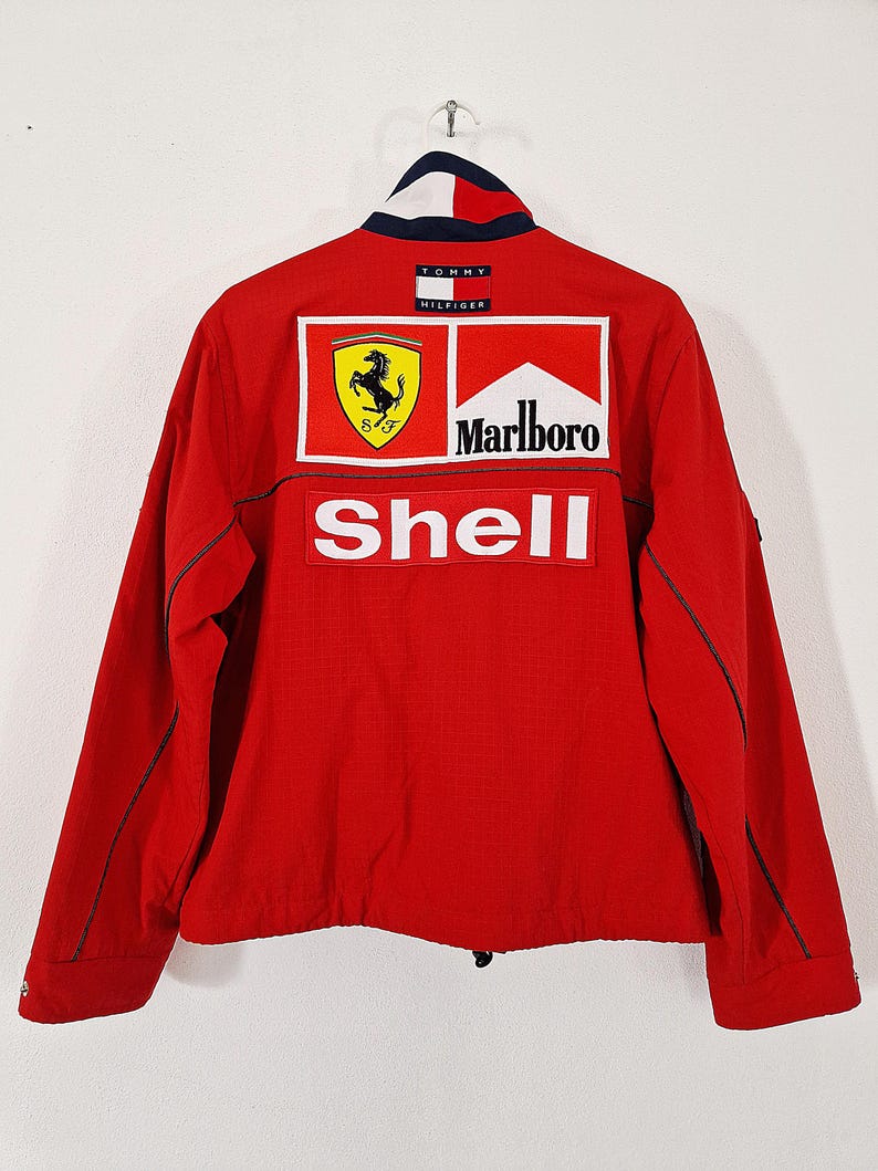 May include: Red jacket with a white, blue, and red collar. The back of the jacket features the Ferrari logo, the Marlboro logo, and the Shell logo. The jacket is a Tommy Hilfiger design.