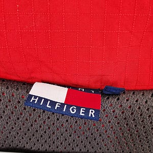 May include: Close-up of a red, quilted fabric. A tag with the word "HILFIGER" in white letters on a navy blue background is visible, along with the brand's logo. The fabric appears to be part of a garment.