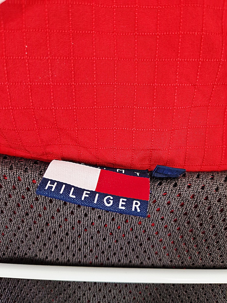 May include: Close-up of a red, quilted fabric. A tag with the word "HILFIGER" in white letters on a navy blue background is visible, along with the brand's logo. The fabric appears to be part of a garment.