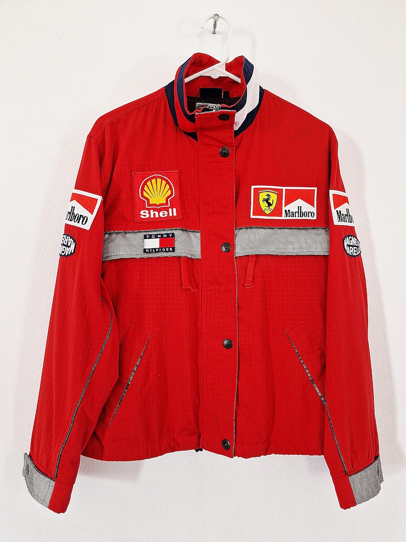 May include: Red racing jacket with multiple sponsor patches, including Shell, Marlboro, and Tommy Hilfiger. The jacket has a stand-up collar with blue and white trim, a gray stripe across the chest, and black buttons.