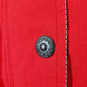 May include: Close-up of a red jacket with a textured, grid-like pattern. A black button with the text "TOMMY HILFIGER" is centered on the jacket. A black and white striped trim runs along the edge of the jacket.