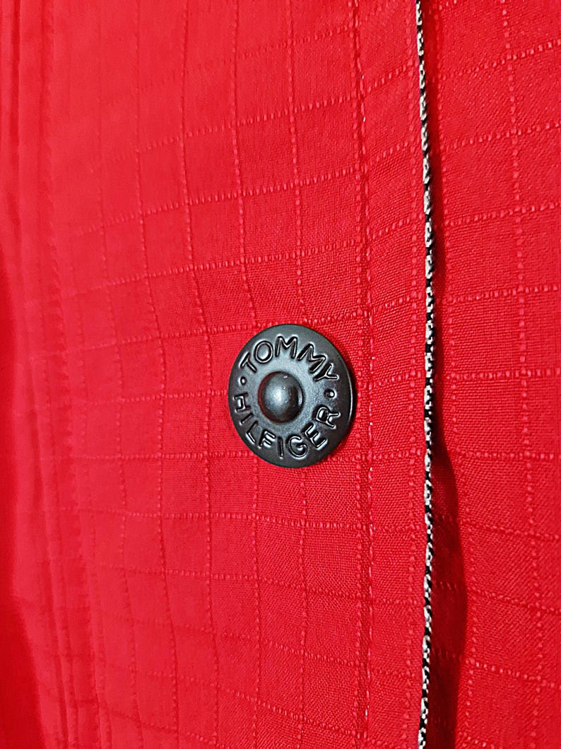 May include: Close-up of a red jacket with a textured, grid-like pattern. A black button with the text "TOMMY HILFIGER" is centered on the jacket. A black and white striped trim runs along the edge of the jacket.