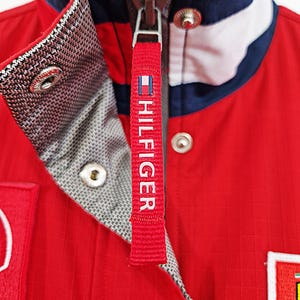 May include: Close-up of a red jacket with a white, navy, and red collar. The jacket has a zipper with a red tag that says "HILFIGER" in white. The jacket has a quilted texture and a gray patterned lining.