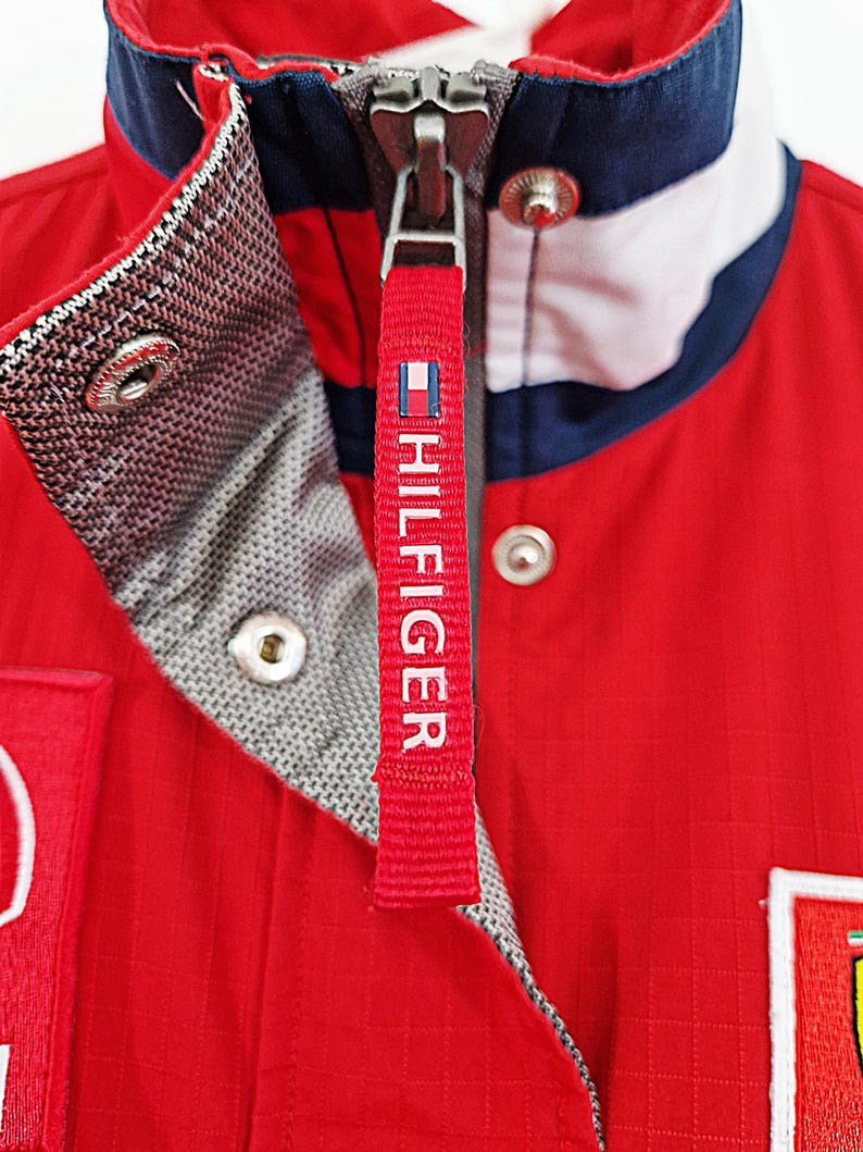 May include: Close-up of a red jacket with a white, navy, and red collar. The jacket has a zipper with a red tag that says "HILFIGER" in white. The jacket has a quilted texture and a gray patterned lining.
