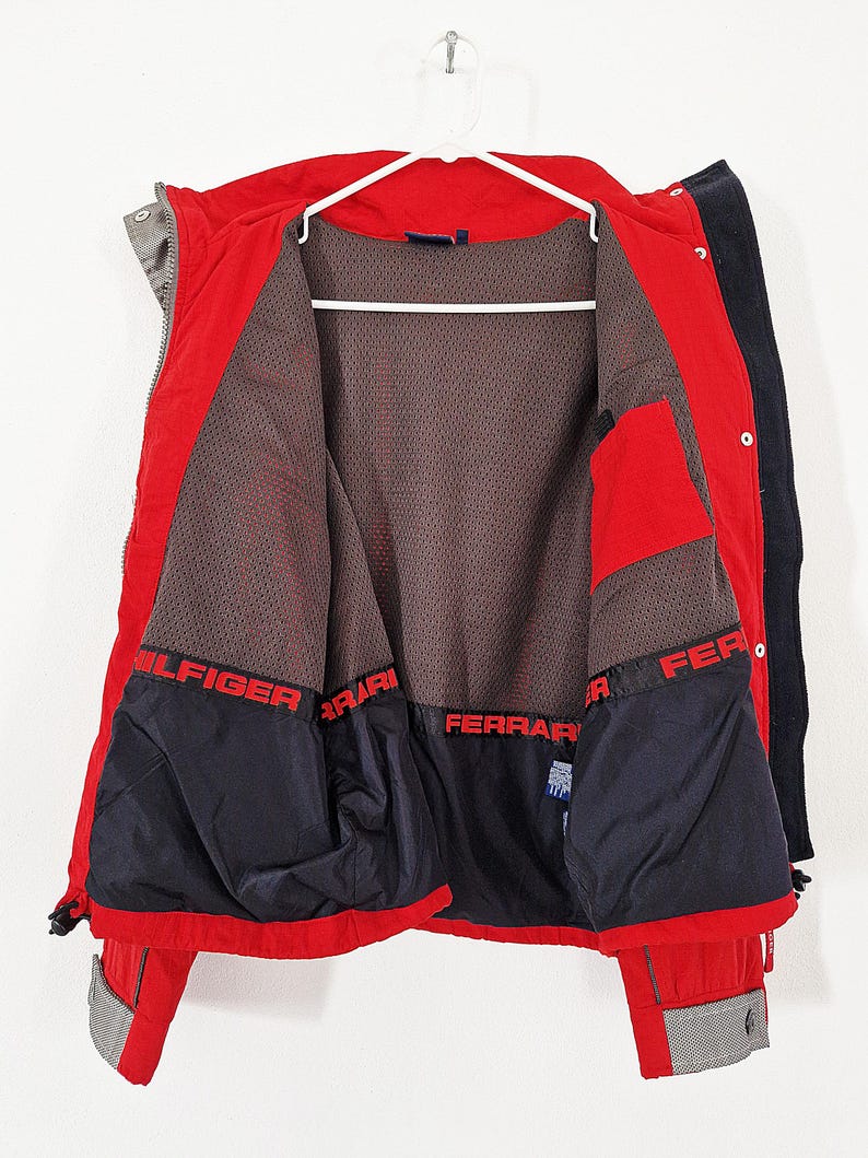 May include: A red and black jacket with the interior visible, hanging on a white hanger. The jacket has the words "HILFIGER" and "FERRARI" in red lettering. The interior is a dark mesh material.