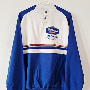 May include: A blue and white long-sleeve pullover sweatshirt featuring a mock neck with two buttons. The chest displays the Rothmans Racing logo and the text Rothmans MITSUBISHI above horizontal red, white, and blue stripes.