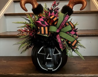 Puede incluir: Un cubo de calabaza de plástico negro relleno de vegetación artificial, flores negras y un gran lazo con varios patrones en verde neón, naranja y rosa. Dos piernas de elfo de color naranja brillante con puños de piel negra sobresalen.