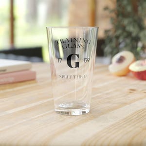 May include: A clear glass with the text "TRAINING GLASS" above a large black "G" and the text "SPLIT THE G" below. The numbers "17" and "59" flank the "G". The glass is on a light wood surface.