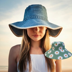 Puede incluir: Una mujer lleva un sombrero de sol de mezclilla azul con un ala ancha. Está de pie frente a una escena de playa. Un segundo sombrero con fondo verde y estampado de margaritas blancas es visible en primer plano.