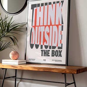 Op de afbeelding: Een ingelijste poster met de woorden "THINK OUTSIDE THE BOX" in vette, witte en rode letters. De poster wordt weergegeven op een houten bank met zwarte metalen poten. Een ronde spiegel en een plant zijn zichtbaar op de achtergrond.