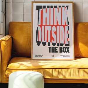 Op de afbeelding: Een ingelijste poster met de woorden "THINK OUTSIDE THE BOX" in vetgedrukte, gestileerde letters. De poster staat in een houten lijst en rust op een gele bank. De tekst is zwart, rood en grijs.