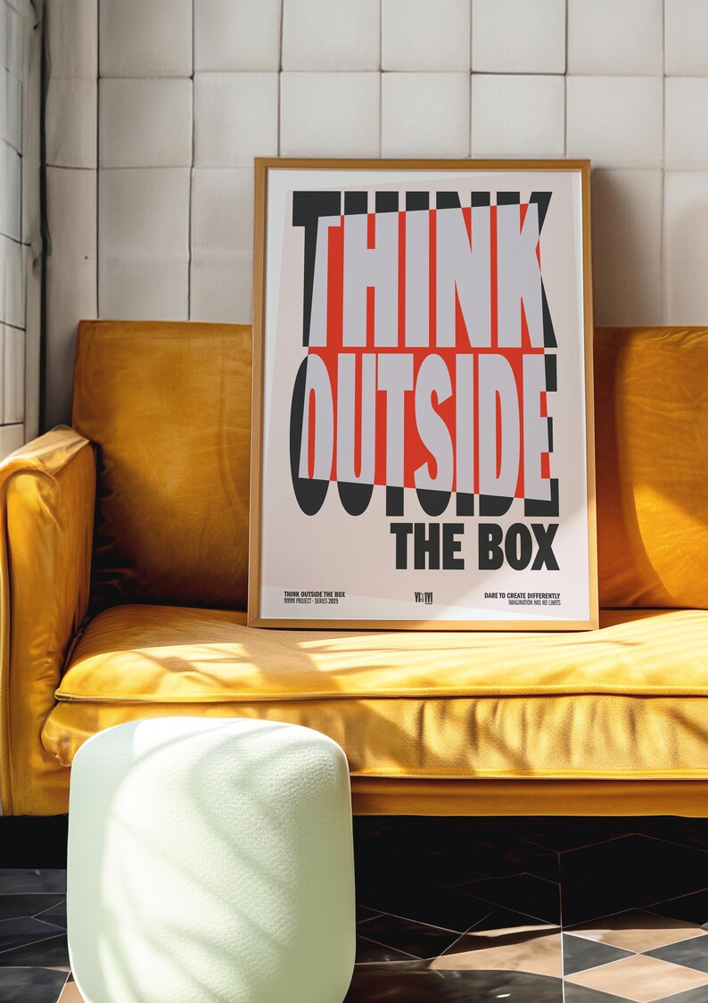 Op de afbeelding: Een ingelijste poster met de woorden "THINK OUTSIDE THE BOX" in vetgedrukte, gestileerde letters. De poster staat in een houten lijst en rust op een gele bank. De tekst is zwart, rood en grijs.