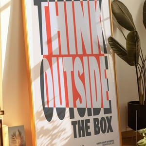 Op de afbeelding: Een ingelijste poster met de tekst "THINK OUTSIDE THE BOX" in een gedurfde, moderne lettertype. De tekst is wit, rood en grijs op een cr&egrave;mekleurige achtergrond. De poster zit in een gouden lijst en staat tegen een muur.
