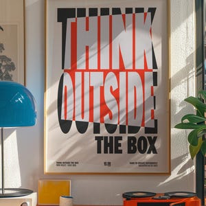 Op de afbeelding: Een ingelijste poster met de tekst "THINK OUTSIDE THE BOX" in een vet, modern lettertype. De poster heeft een cr&egrave;mekleurige achtergrond met zwarte en rode letters. De afbeelding toont ook een blauwe lamp, een platenspeler en andere decoratieve items.