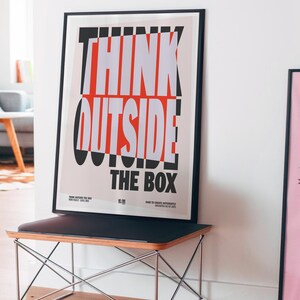 Op de afbeelding: Een ingelijste poster met de woorden "THINK OUTSIDE THE BOX" in dikke witte en rode letters. De poster wordt weergegeven op een zwarte bank met een houten blad en een metalen frame. De kamer heeft een modern design.