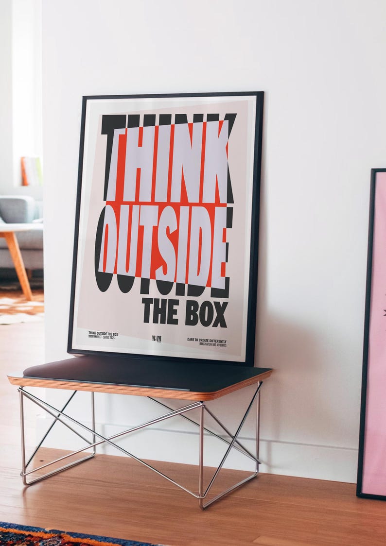 Op de afbeelding: Een ingelijste poster met de woorden "THINK OUTSIDE THE BOX" in dikke witte en rode letters. De poster wordt weergegeven op een zwarte bank met een houten blad en een metalen frame. De kamer heeft een modern design.