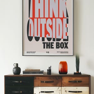 Op de afbeelding: Een ingelijste poster met de woorden "THINK OUTSIDE THE BOX" in vetgedrukte, gestileerde letters. Boven een houten dressoir met diverse decoratieve items, waaronder een vaas en een kleine plant. Het dressoir heeft lades in zwart, wit en houttinten.