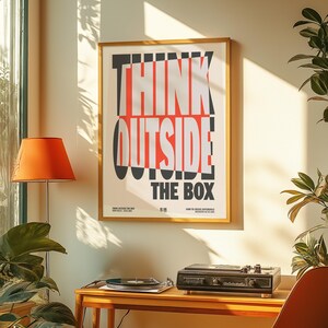 Puede incluir: Un póster enmarcado con las palabras "THINK OUTSIDE THE BOX" en un diseño moderno y audaz. El texto es en negro, rojo y blanco sobre un fondo crema. El póster está colgado en una pared, con un tocadiscos y una lámpara en primer plano.
