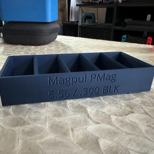 May include: A dark blue plastic magazine holder with five compartments. The holder has the text "Magpul PMag 5.56 / .300 BLK" printed on the front.