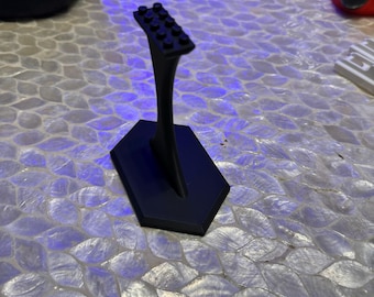 May include: A black plastic stand with a hexagonal base and a curved stem. The top of the stand is a flat surface with a row of small black squares.